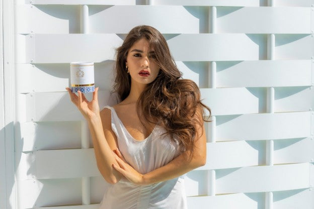 A woman with long wavy brown hair stands against a white slatted wall, holding a small cylindrical container with a white label and blue patterned base.