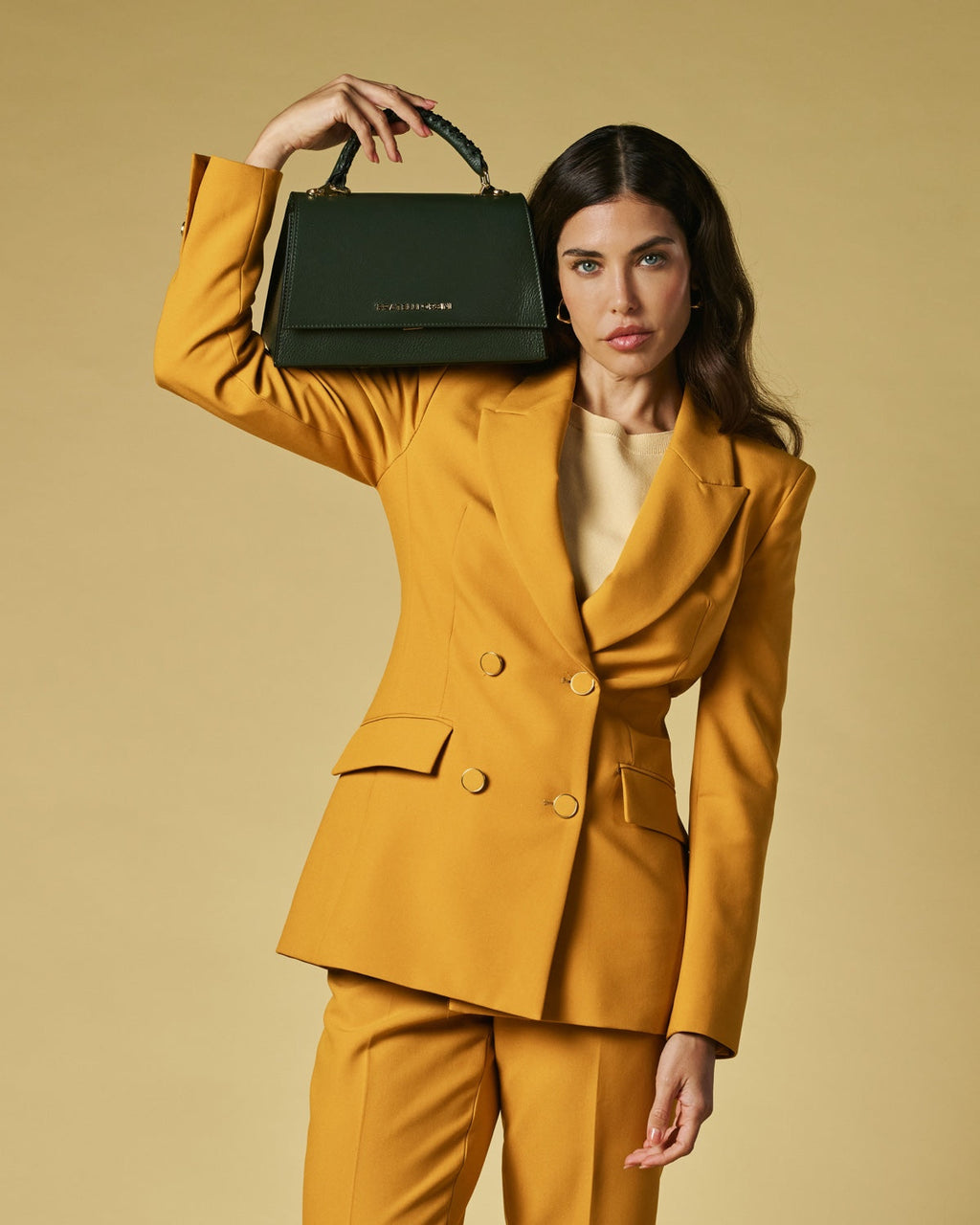 Woman in a mustard yellow suit holding a green handbag against a beige background