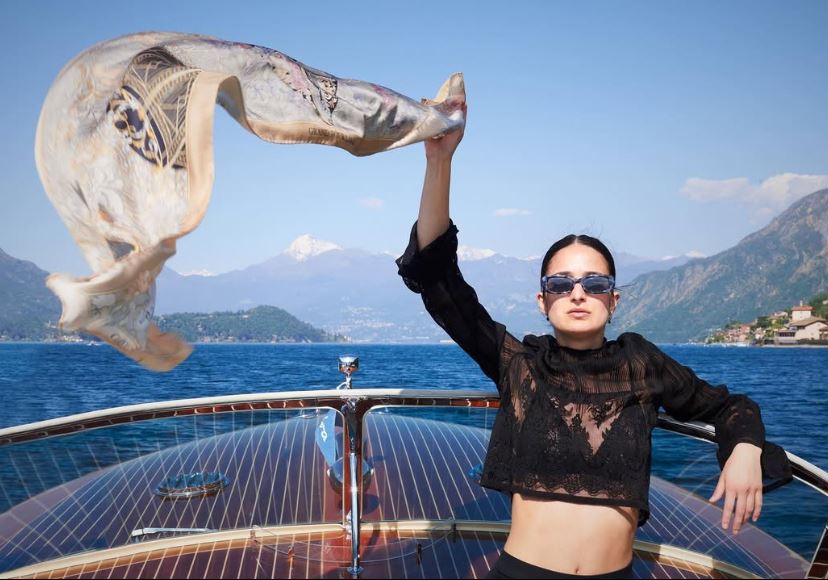 woman with black shirt and sunglasses, waving a golden silk scarf in the air. She's sitting in a motorboat on lake Como. On the background the lake's water, Lenno, Tremezzina, Villa Balbianello and the mountains