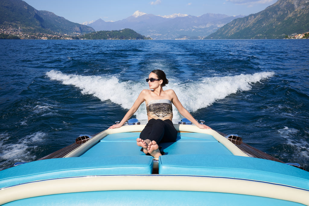 woman wearing sunglusses and a silk scarf as shirt, sitting on a motorboat on the waters of Lake Como. On the background the lake, Tremezzina, Lenno, Villa Balbianello and the mountains
