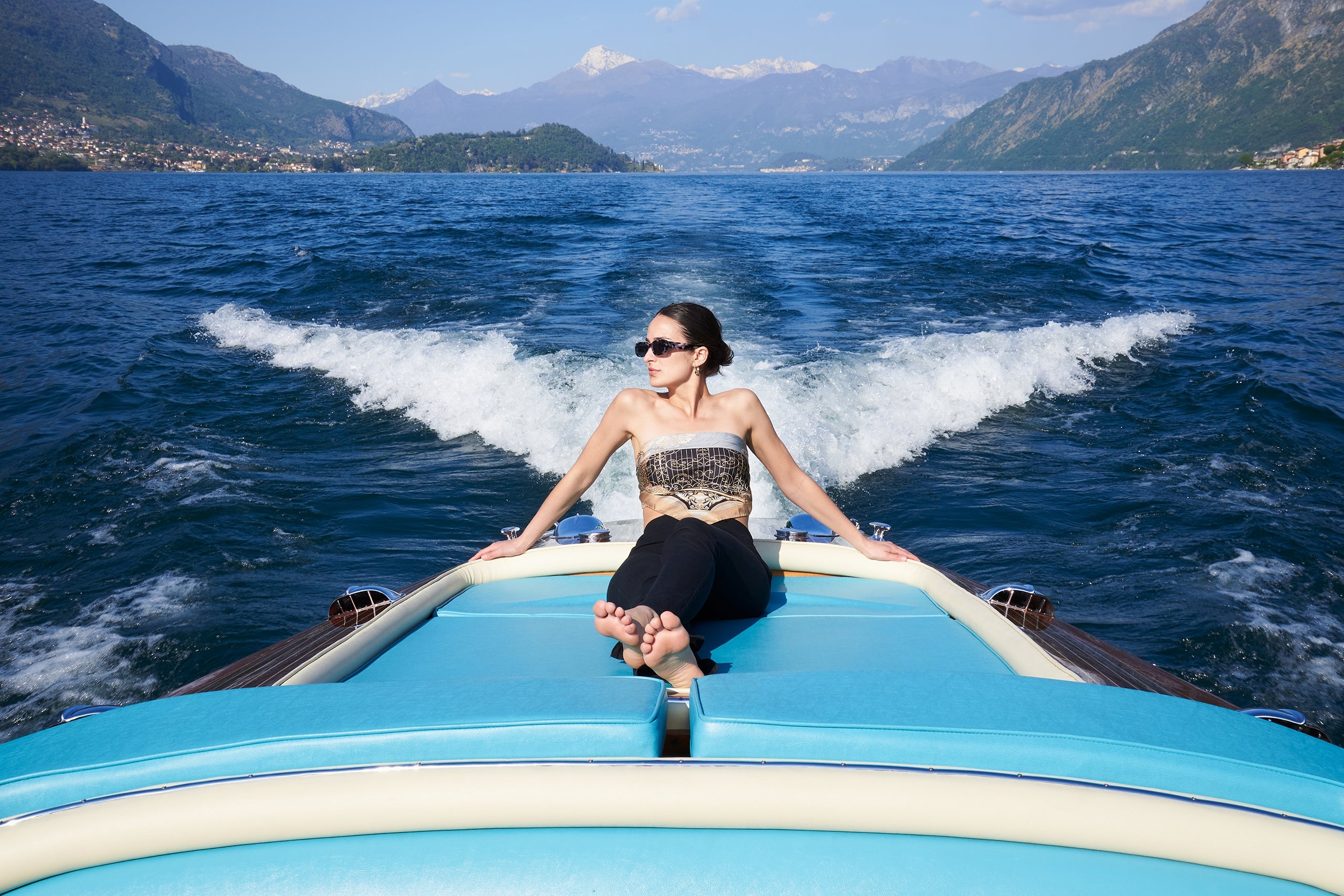 woman wearing sunglusses and a silk scarf as shirt, sitting on a motorboat on the waters of Lake Como. On the background the lake, Tremezzina, Lenno, Villa Balbianello and the mountains