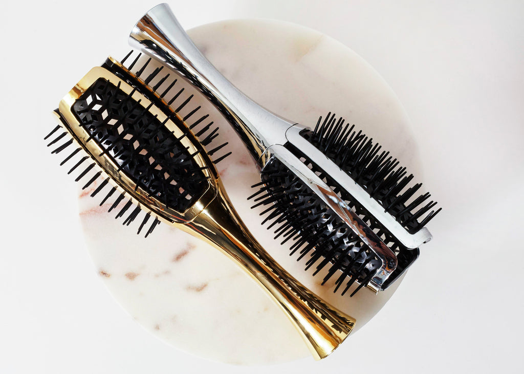 Two hairbrushes with black bristles, one featuring a gold metallic handle and the other a silver metallic handle, displayed on a round white marble coaster against a white background.