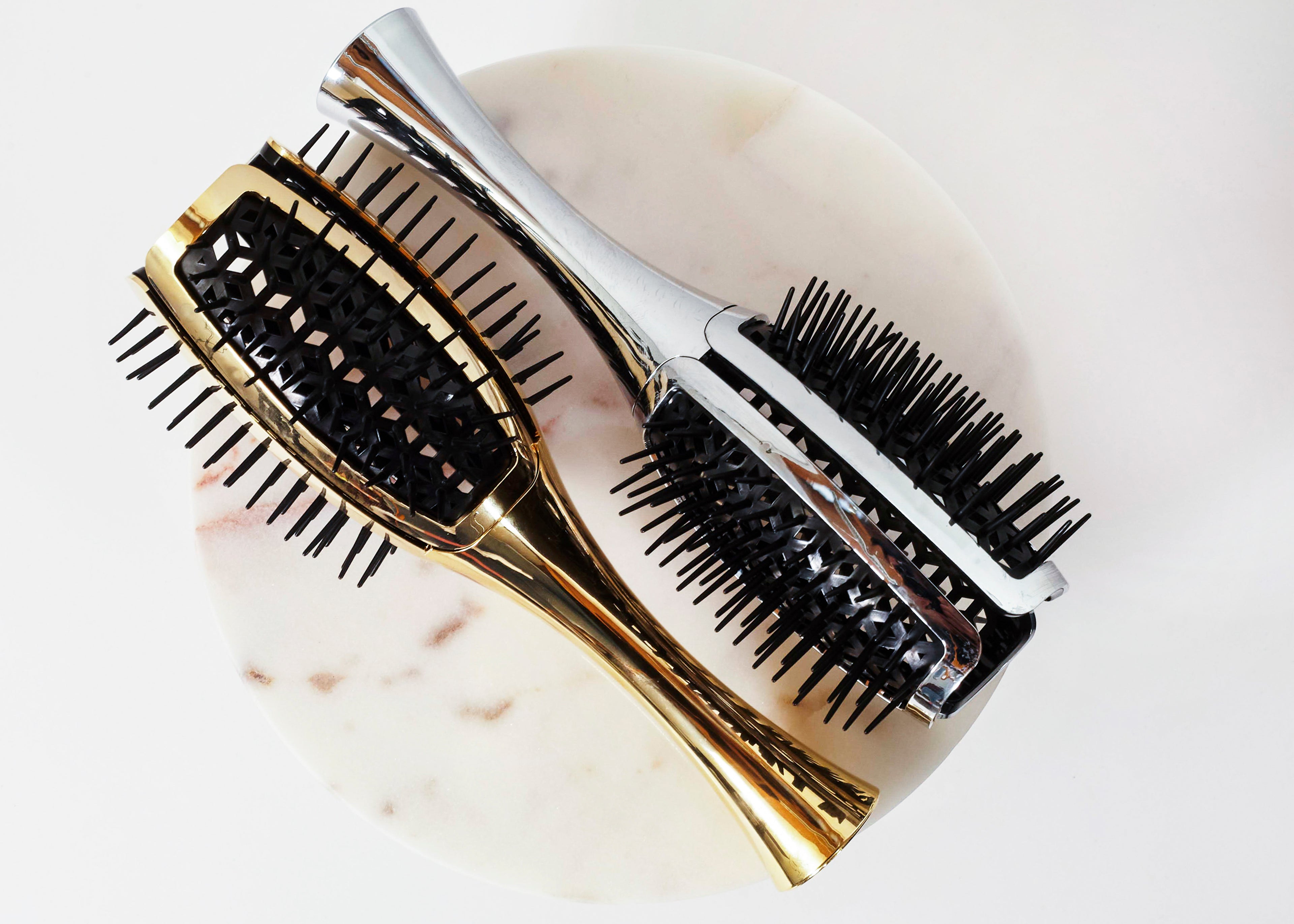 Two hairbrushes with black bristles, one featuring a gold metallic handle and the other a silver metallic handle, displayed on a round white marble coaster against a white background.