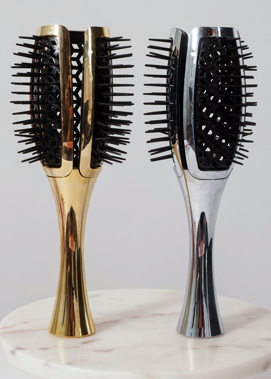 Two paddle hairbrushes standing upright on a white marble surface against a light gray background. One brush has a polished gold handle and black bristles, and the other has a polished silver handle with black bristles.
