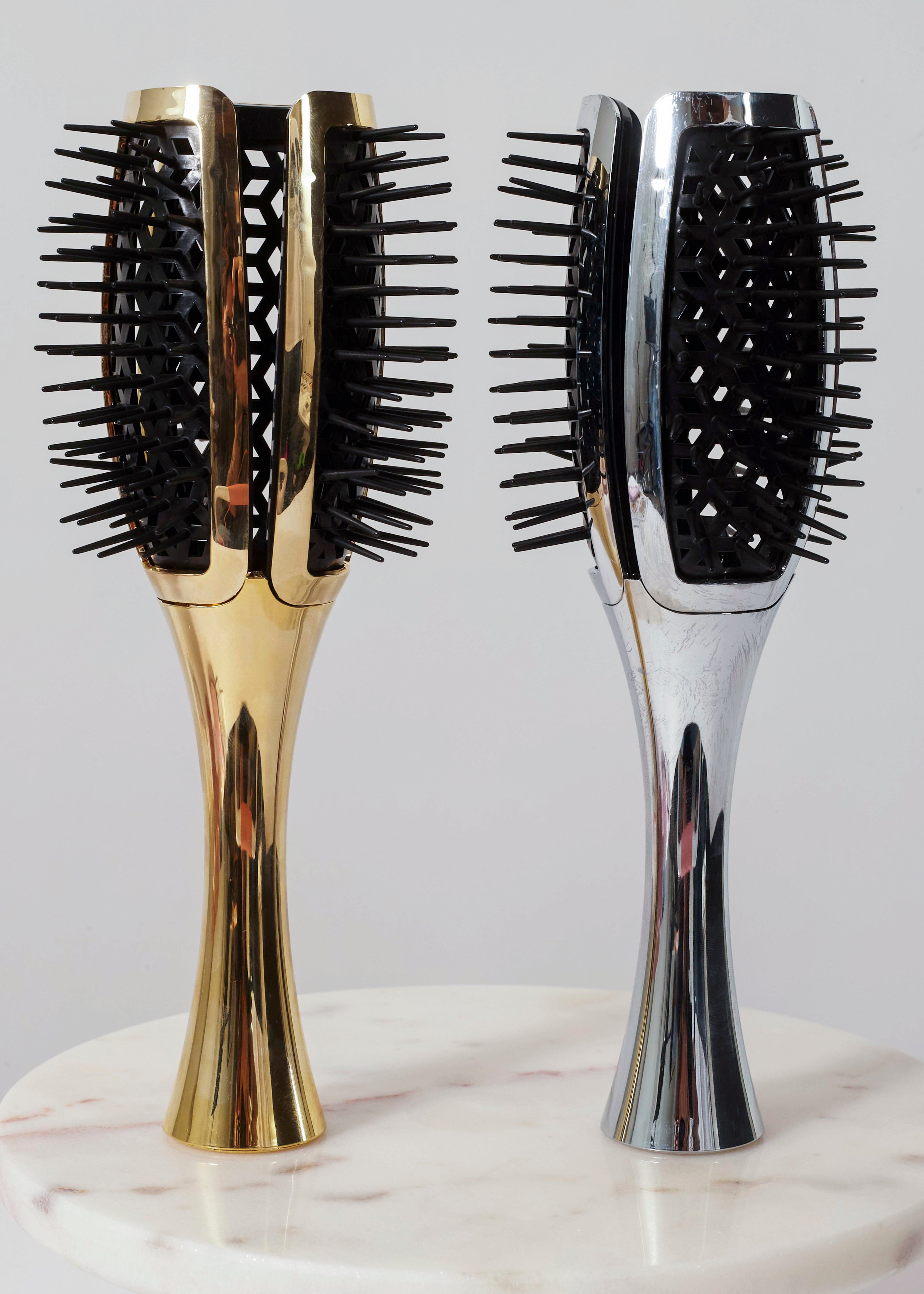 Two paddle hairbrushes standing upright on a white marble surface against a light gray background. One brush has a polished gold handle and black bristles, and the other has a polished silver handle with black bristles.
