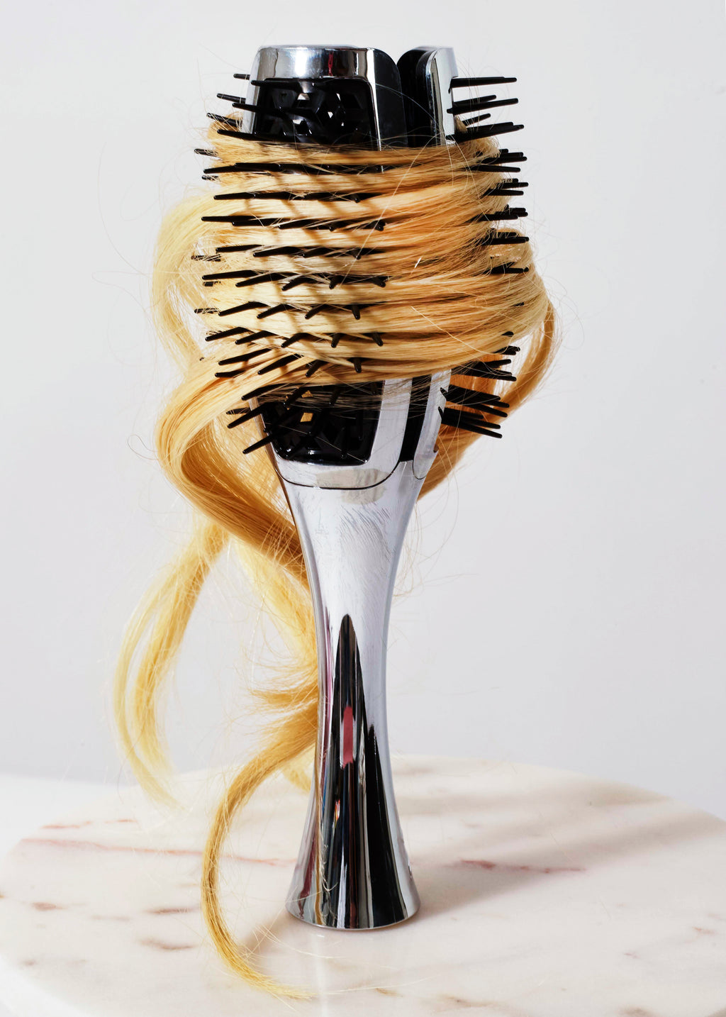 A round hairbrush with a shiny silver handle and black bristles, wrapped in a loop of blonde hair, standing on a light marble surface against a white background.