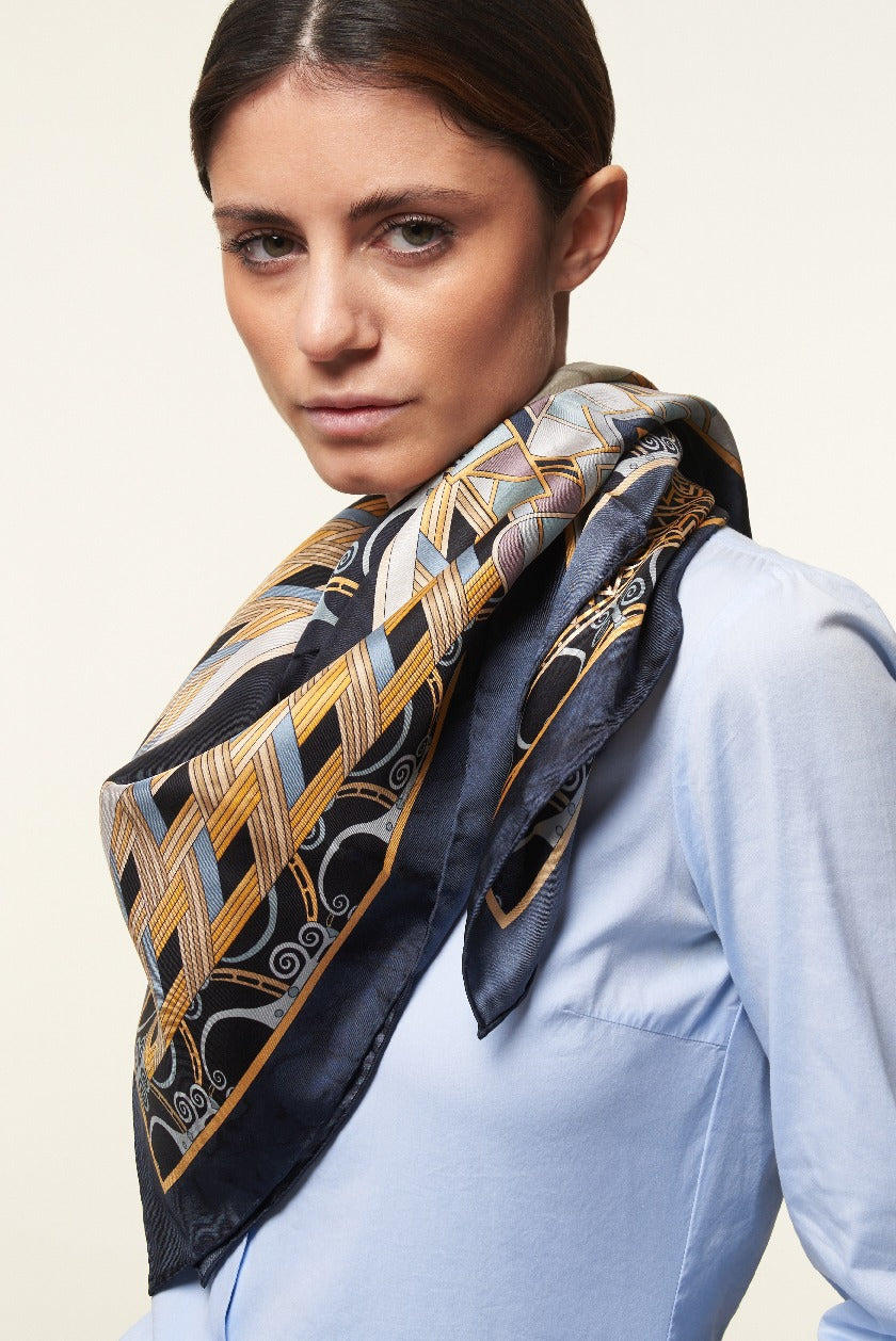brunette woman wearing a white shirt and a black, gold and white silk scarf with geometric patterns