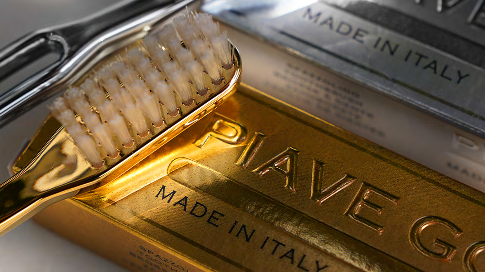 A close-up of a gold toothbrush with white bristles resting on a shiny gold box embossed with the brand name 'PIAVE' and 'MADE IN ITALY'.
