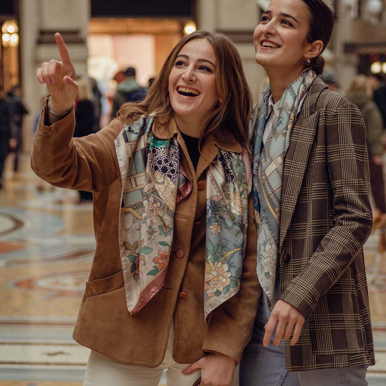 two women laughing inside galleria Vittorio Emanuele in Milan, wearing silk scarves around their neck