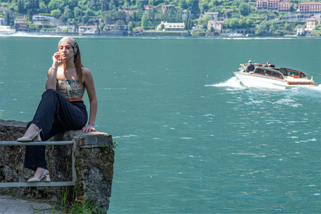 woman wearing a silk bandana and a silk scarf as shirt. On the background a motorboat on the waters of lake Como, and a lakeside town