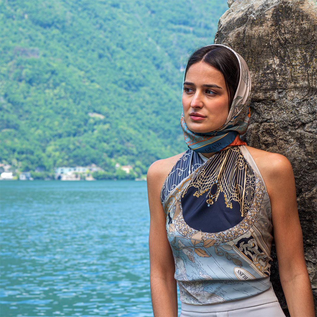 woman dressed with a blue silk scarf, with another one wrapped around her head. On the background the water of Lake Como and its mountains