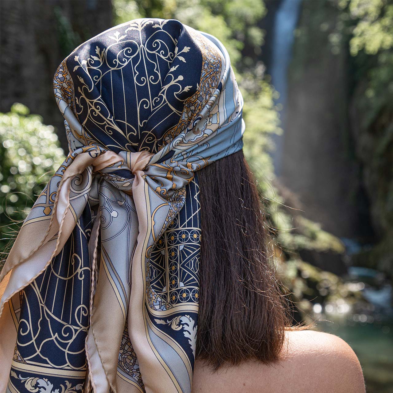 woman wearing a black and gold silk scarf on her head. On the background a waterfall in Nesso, on lake Como