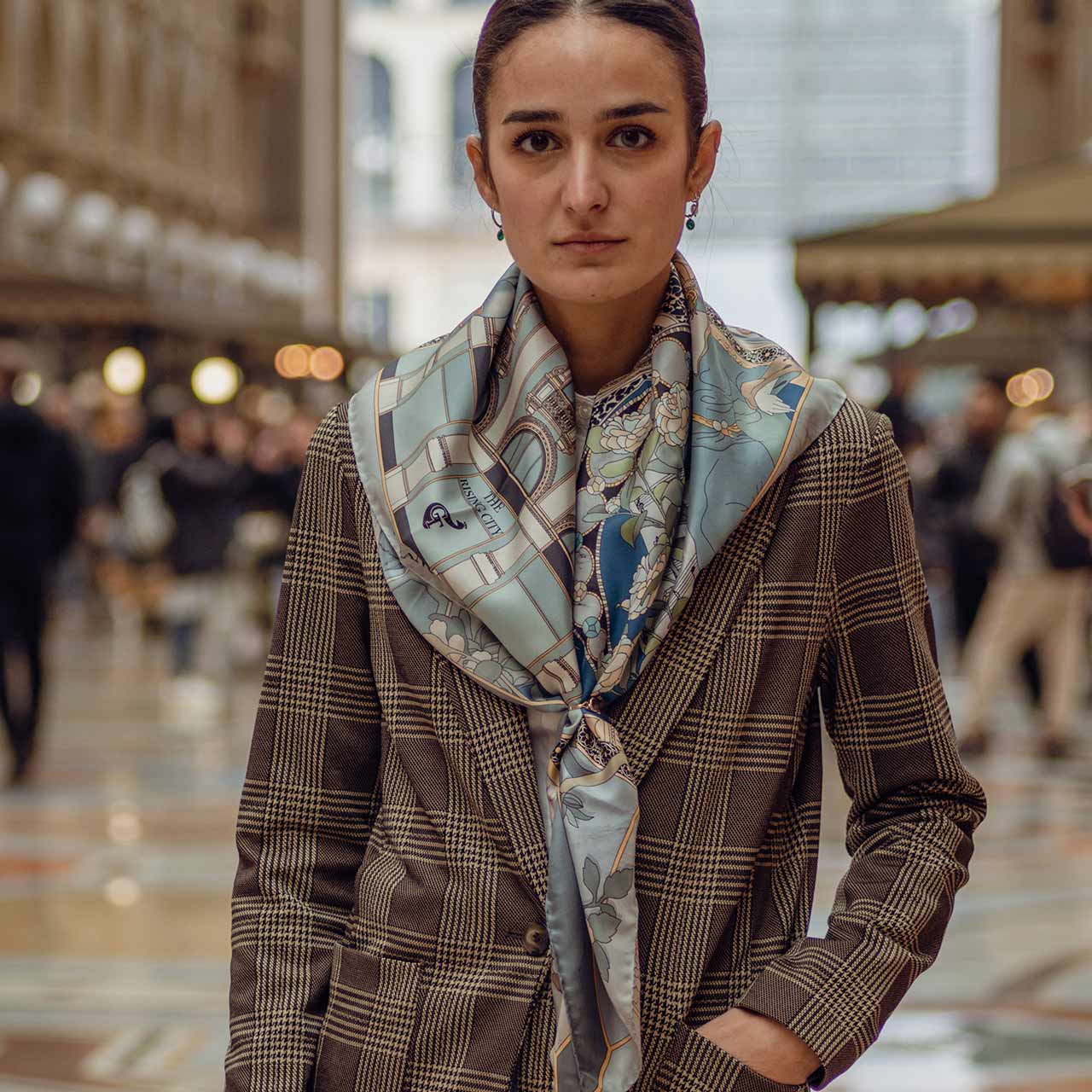 woman wearing a brown coat and blue silk scarf. On the background galleria Vittorio Emanuele in Milan